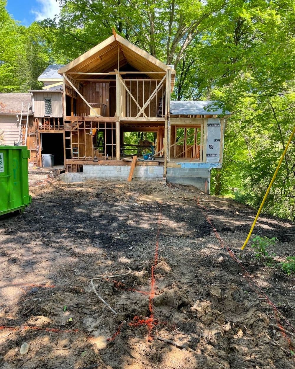 House under construction with exposed framing, surrounded by trees and construction equipment.