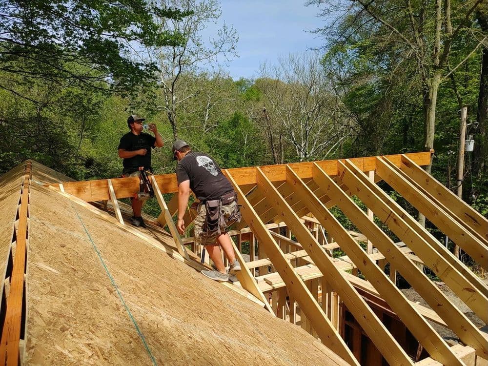 Two workers framing a roof on a wooden structure in a forested area.
