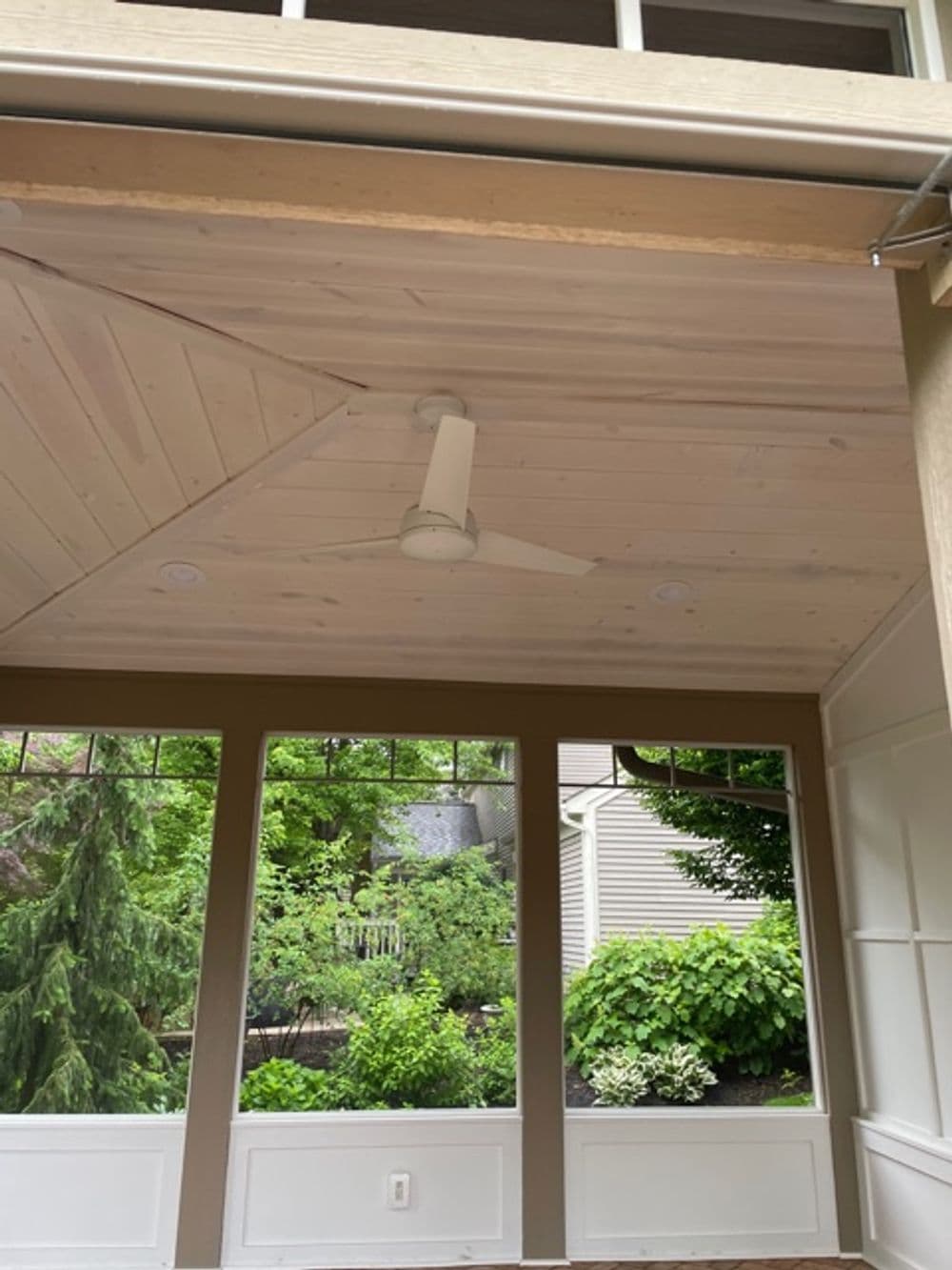 Interior view of a covered porch with a ceiling fan and large windows overlooking greenery.