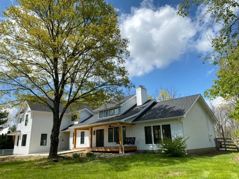 Modern white house with a porch, large yard, and trees under a blue sky with clouds.