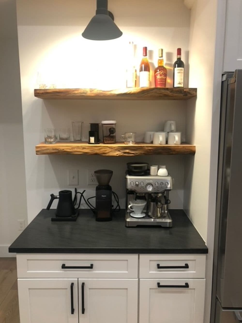 Modern kitchen coffee station with black countertop, coffee machine, shelves, and decorative bottles.